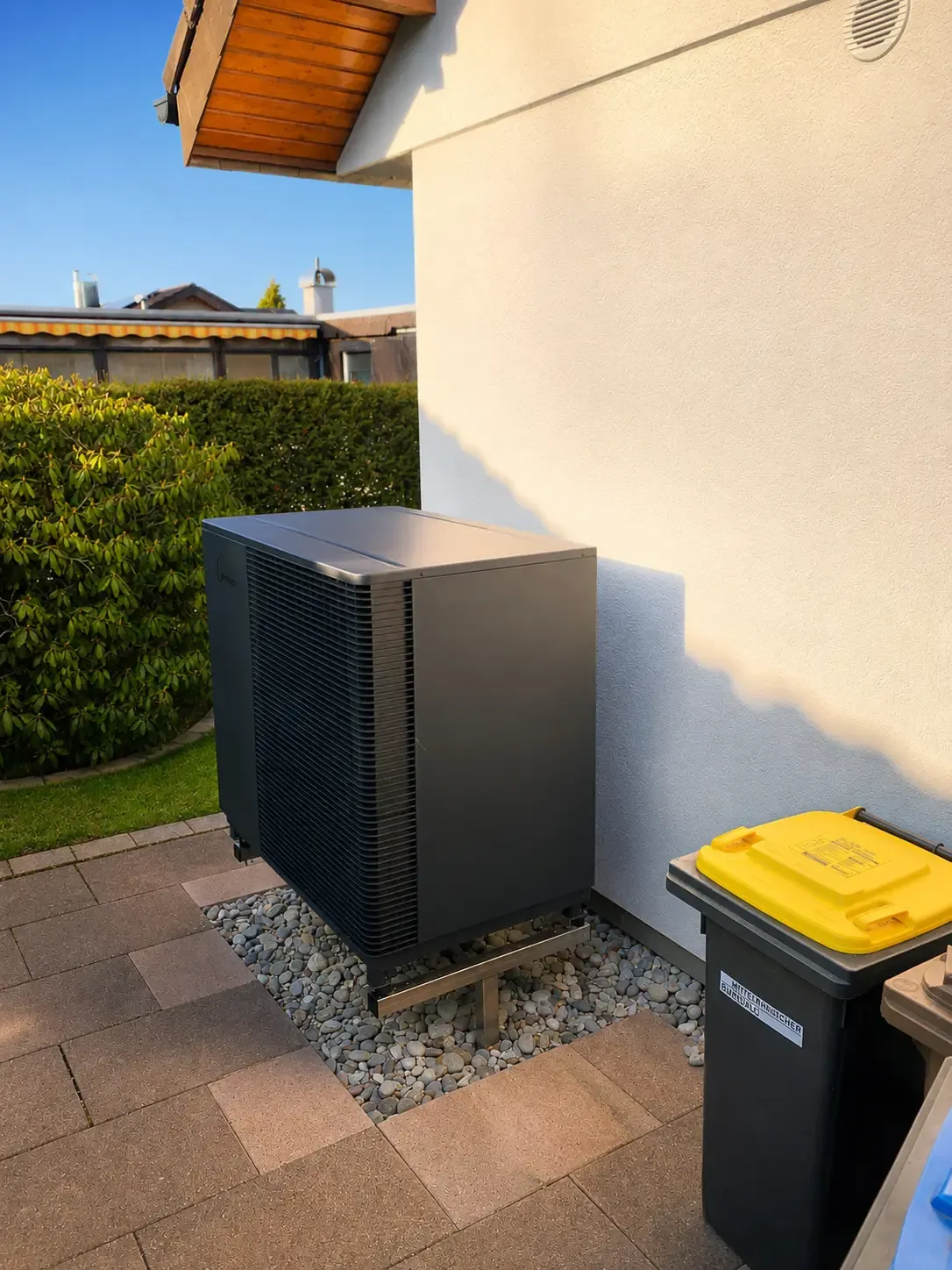 Außeneinheit einer modernen Wärmepumpe an der Hausfassade mit gepflastertem Gehweg und gepflegter Gartenanlage. Die Heizlösung nutzt erneuerbare Energie aus der Umgebung und unterstützt eine nachhaltige Wärmeversorgung.
