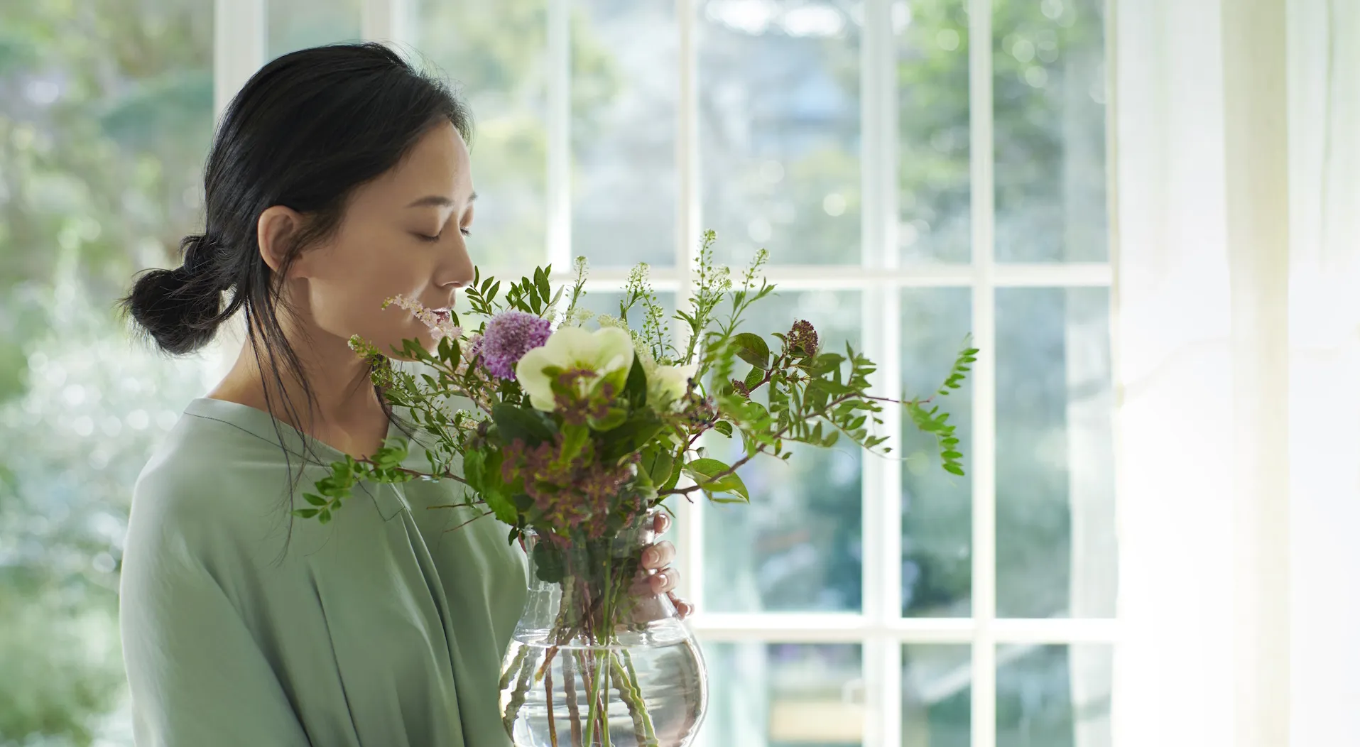 Japanische Frau steht vor einem großen Fenster und hält eine Vase mit wohlduftenden Blumen in der Hand.