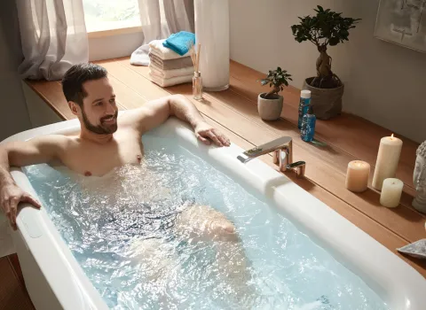 Der Mann liegt entspannt in einer Badewanne von Mauersberger mit sprudelndem Wasser, während Kerzen und eine Zimmerpflanze auf der hölzernen Wannenablage eine ruhige Stimmung schaffen. 
