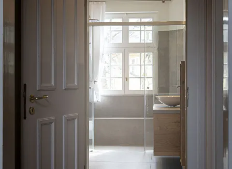 Eine stilvolle Jugendstil-Tür mit weißem Rahmen und klassischen Kassettendetails öffnet sich zu einem lichtdurchfluteten Badezimmer.