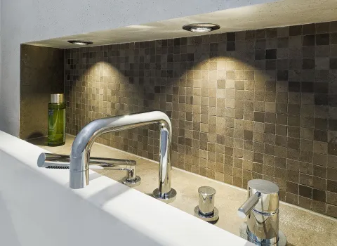 Unterputz-Badewannenarmatur mit glänzendem Chromfinish vor einer Wand mit kleinen Mosaikfliesen in Erdtönen.