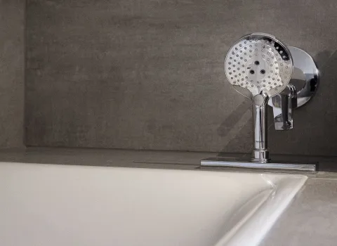 Eine moderne Badewannenarmatur mit einer verchromten Handbrause ist an einer dunkelgrauen Betonwand montiert.