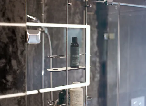 Detailaufnahme einer Dusche in dessen Glaswand sich das umlaufende Licht des rechteckigen Waschtisch-Spiegels spiegelt.