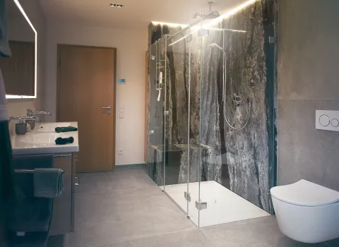 Badezimmer mit großer Glasdusche, edler Natursteinwand, stilvollem Waschtisch und warmen Lichtkonzept.