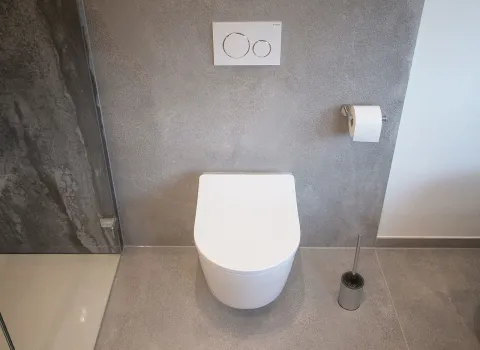 Weißes, modernes WC neben Dusche, vor grauer Wand aus Großformatfließen.
