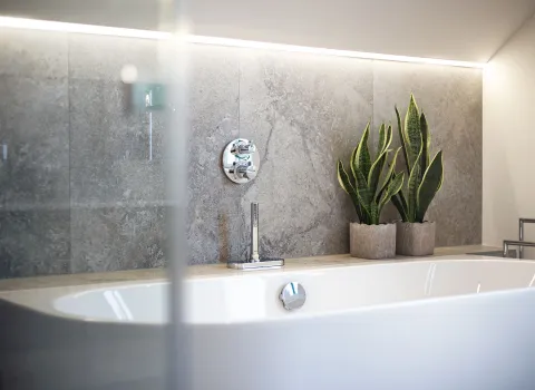 Eine wandstehende Badewanne ist vor einer mit Natursteinfliesen verkleideten Wand positioniert.