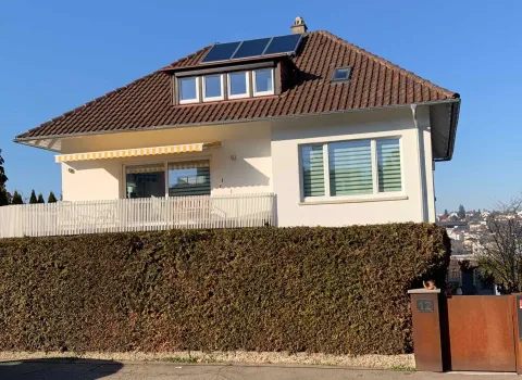 Ein modernes Einfamilienhaus mit cremefarbener Fassade und braunem Ziegeldach steht unter einem klaren blauen Himmel.