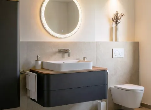 Fotorealistischer Planungsentwurf eines kompakten Comfort-Badezimmers mit Waschtisch, rundem Leuchtspiegel und schwebender Toilette in gedimmten Licht.