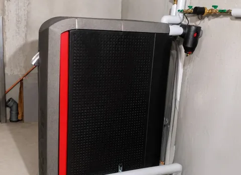 Ein moderner Pelletkessel mit schwarzer Front und roten Designelementen ist in einem Technikraum installiert. Die Heizungsanlage ist an sauber gedämmte Rohrleitungen angeschlossen und sorgt für eine effiziente Wärmeversorgung des Gebäudes. Die professionelle Installation unterstreicht eine nachhaltige und energieeffiziente Heizlösung.