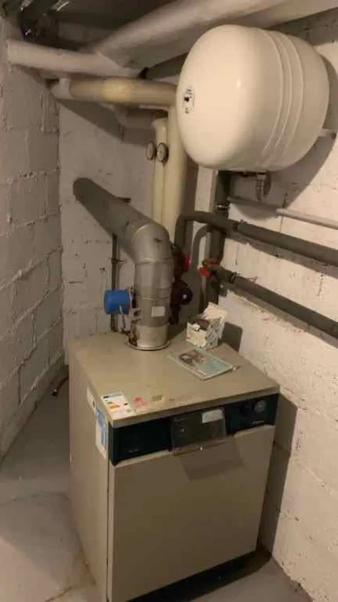 Das Bild zeigt eine alte Heizungsanlage in einem Kellerraum mit weiß verputzten Wänden. 