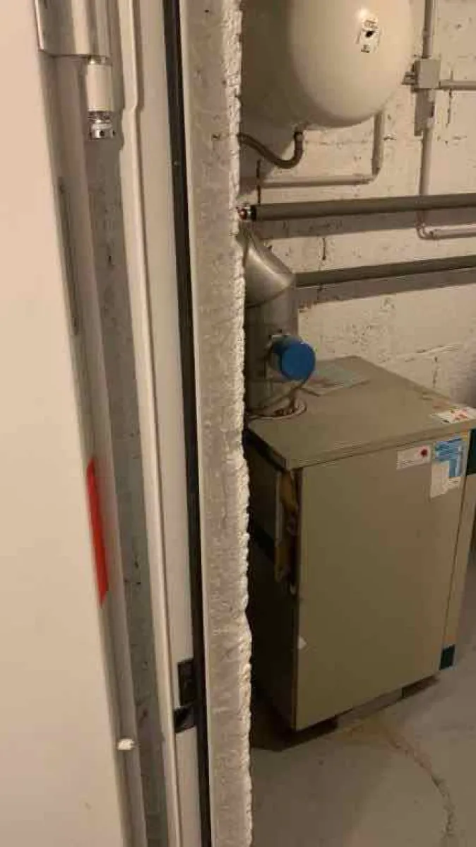 Durch eine geöffnete Tür ist eine alte Heizungsanlage im Keller sichtbar, bestehend aus einem kompakten Heizkessel mit Rohrleitungen und einem weißen Ausdehnungsgefäß.