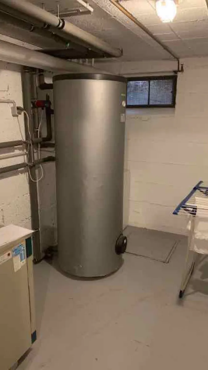 Ein silberner Warmwasserspeicher steht in einem hell gestrichenen Kellerraum neben einer alten Heizungsanlage. 