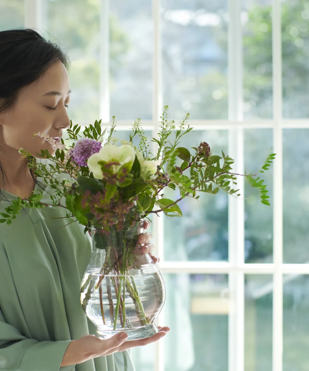Japanische Frau steht vor einem großen Fenster und hält eine Vase mit wohlduftenden Blumen in der Hand.