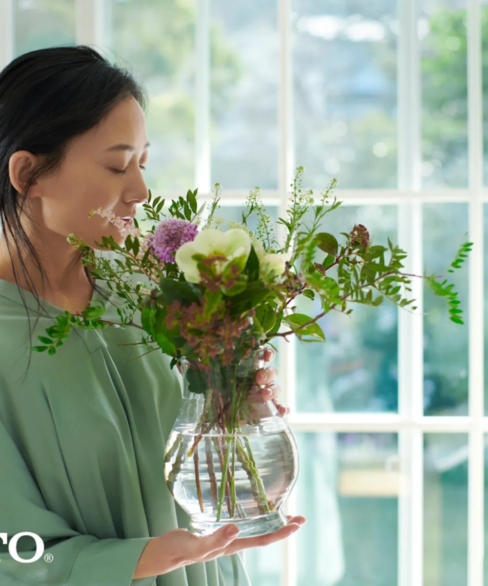 Japanische Frau steht vor einem großen Fenster und hält eine Vase mit wohlduftenden Blumen in der Hand.