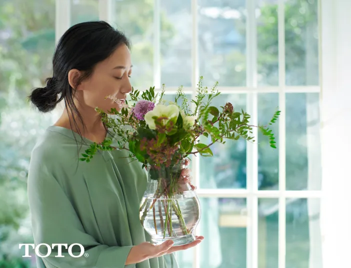 Japanische Frau steht vor einem großen Fenster und hält eine Vase mit wohlduftenden Blumen in der Hand.